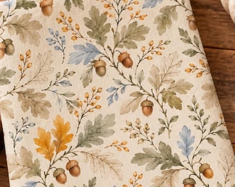 May include: A rectangular kitchen towel featuring a repeating pattern of oak leaves, acorns, and small sprigs in shades of muted green, golden yellow, and soft blue on a cream-colored background, folded neatly into a vertical orientation.