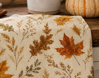 May include: A cream-colored kitchen towel featuring a repeating pattern of autumn leaves, including maple and oak leaves in shades of orange, brown, and muted green, along with delicate sprigs of dried grasses and small berries.