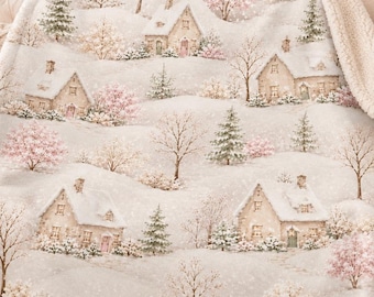 May include: A plush throw blanket featuring a winter village scene with snow-covered houses, evergreen trees, and bare trees with soft pink accents. The blanket has a cream-colored sherpa fleece backing and a folded edge showing the texture.
