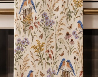 May include: A rectangular kitchen towel featuring a cream-colored background with a repeating pattern of bluebirds perched on wooden posts, surrounded by various wildflowers, tall grasses, and small brown butterflies in shades of blue, yellow, and green.