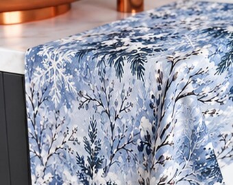 May include: A rectangular fabric runner featuring a winter scene with blue and white snowflakes, snow-covered pine trees, and bare branches. The runner is draped over a dark cabinet edge near a copper-colored basin and faucet fixture.