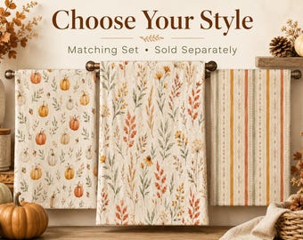May include: Three rectangular kitchen towels hanging on a rod. The left towel features a pumpkin and leaf pattern. The center towel displays a botanical print of wildflowers and grasses. The right towel has vertical stripes in orange and green.