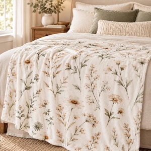 Field Sage and Cream plush blanket styled across the foot of a neutral bed, showing the soft botanical floral print in a warm, cozy bedroom setting.