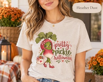 May include: A heather dust colored t-shirt featuring a graphic of a smiling radish character with green leaves, wearing red shoes and a bow, holding a small bag. The text reads pretty pickle halloween in red and green script and block lettering.