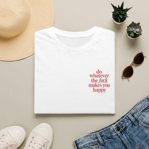 Op de afbeelding: Een wit t-shirt met de rode tekst 'do whatever the fuck makes you happy' gedrukt aan de rechteronderkant. Het t-shirt heeft een ronde hals en korte mouwen, gepresenteerd in een platte compositie met een strooien hoed en een zonnebril.