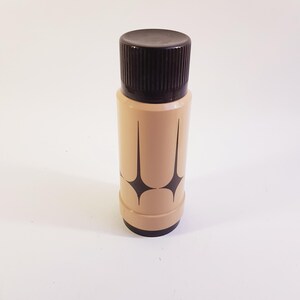 Op de afbeelding: Een beige fles met een donkerbruine dop. De fles heeft een geometrisch sterrenpatroon in donkerbruin. De cilindrische fles lijkt van plastic te zijn.