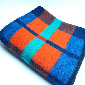 May include: A folded, rectangular blanket with a vibrant, geometric pattern. The design features large squares in shades of orange, teal, and blue, separated by dark blue lines. A thin red line accents the center. The blanket has a soft, textured appearance.