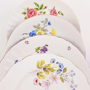 Op de afbeelding: Een stapel van vier witte keramische borden met geschulpte randen, elk voorzien van verschillende bloemmotieven. De ontwerpen bevatten roze rozen, blauwe en gele bloesems, paarse viooltjes en een mix van wilde bloemen.
