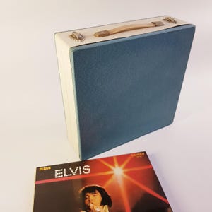 Pode incluir: Uma mala de discos azul vintage com parte superior e alça creme. A mala é retangular e possui fechos de metal. Um álbum do Elvis com o logotipo da RCA está na frente.