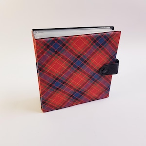 May include: A red, blue, and black plaid photo album with a black leather strap closure. The album is square-shaped and has a black spine. The plaid pattern is a classic design.
