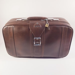 Può includere: Una valigia in pelle marrone con maniglia superiore e hardware color argento. La valigia ha una chiusura con fibbia e un piccolo lucchetto con chiavi attaccate. Il bagaglio è rettangolare e sembra vintage.