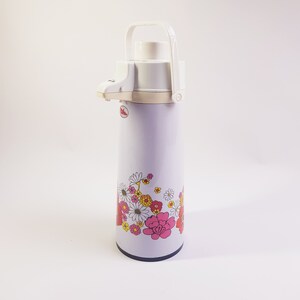 Op de afbeelding: Een witte en paarse thermoskan met een bloemenpatroon. De kan heeft een witte handgreep en een witte tuit. De bloemen zijn roze, rood, geel en wit.
