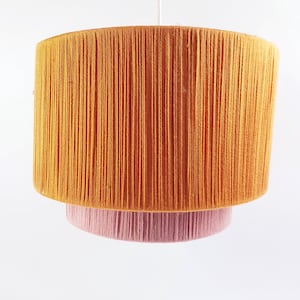 Op de afbeelding: Een hanglamp met twee niveaus. De bovenste laag is cilindrisch en oranje, de onderste laag is een kleinere, cirkelvormige vorm in roze. De lamp is gemaakt van garen of draad, waardoor een textuur ontstaat. Opgehangen aan een witte kabel.