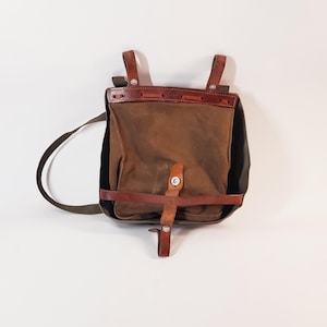 Puede incluir: Mochila de lona marrón y cuero con cierre de solapa y correas ajustables. La bolsa presenta un cuerpo de lona marrón oscuro, complementado con correas y detalles de cuero. El diseño incluye una correa para el hombro y un cierre de hebilla.