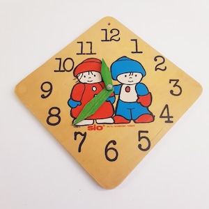 Op de afbeelding: Een vierkante houten klok met een gele wijzerplaat en zwarte cijfers. De klok heeft een groene wijzer en twee cartoonkinderen in rood en blauw. De tekst "sio by, amsterdam, holland" staat op de wijzerplaat van de klok.