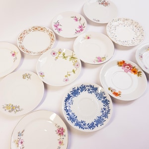 Op de afbeelding: Een verzameling vintage witte porseleinen schoteltjes met bloemmotieven. De schoteltjes hebben verschillende patronen, waaronder roze, oranje en blauwe bloemen, en gouden randen. De schoteltjes zijn gerangschikt op een wit oppervlak.