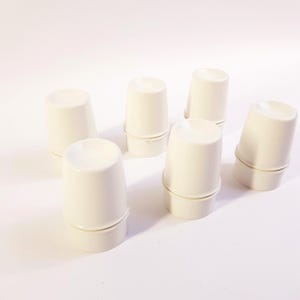 Peut inclure: Six gobelets coniques blanc cassé sont disposés sur une surface blanche. Chaque gobelet a une base légèrement évasée et un dessus lisse et arrondi. Les gobelets sont espacés uniformément, certains étant légèrement inclinés.