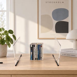 May include: A pair of clear acrylic bookends with black metal bases, positioned on a wooden surface to support a row of compact discs. The transparent vertical panels provide a clean, unobtrusive support system for organizing media collections.