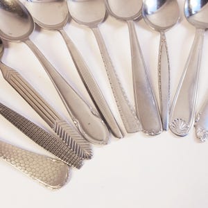 Può includere: Un assortimento di cucchiai d'argento vintage con vari disegni del manico, tra cui motivi geometrici, linee e un motivo a conchiglia. I cucchiai sono disposti su una superficie bianca, mettendo in mostra le loro superfici lucide e gli stili unici.