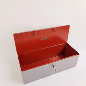 May include: A rectangular metal cash box with a textured light gray exterior and a vibrant red interior. The lid is open, revealing a small oval metal emblem on the inside. A silver-colored key is inserted into the front keyhole lock mechanism.
