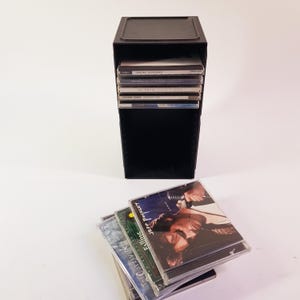 May include: A black CD storage tower with a stack of CDs inside. Several CDs are stacked in front of the tower. The CDs have various album art and titles. The tower is rectangular and has a lid.