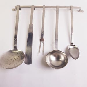 Op de afbeelding: Een set roestvrijstalen keukengerei dat aan een metalen rek hangt. De set bevat een schuimspaan, spatel, vork, pollepel en lepel. Elk keukengerei heeft een lange steel en een decoratief element aan de bovenkant.