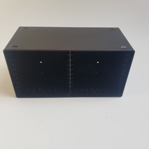 May include: Black plastic storage box with two compartments and a divider. The box is rectangular and measures approximately 30 cm in length.