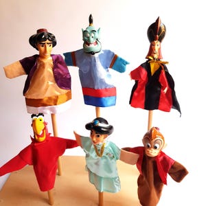 Op de afbeelding: Een set van zes handpoppen op houten stokjes, met personages uit Aladdin, waaronder een man in een paars vest, een blauwe geest, een man in een zwart met rood gewaad, een rode papegaai, een vrouw in een lichtblauwe jurk en een bruine aap.