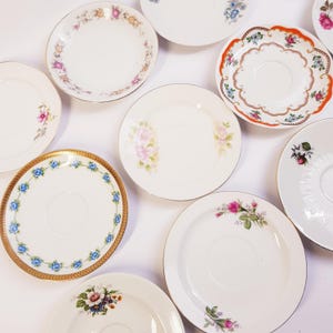 Op de afbeelding: Een verzameling vintage porseleinen schotels en kleine borden. De borden hebben bloemmotieven in verschillende kleuren, waaronder roze, blauw en oranje, met gouden randen. Sommige hebben gedetailleerde ontwerpen, andere eenvoudigere motieven.
