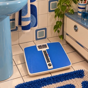May include: A square mechanical bathroom scale with a bright blue top surface and a white central display panel. The scale features a vertical weight gauge with black numbering and a red indicator needle, designed for measuring body weight in kilograms.