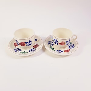 Könnte beinhalten: Zwei weiße Teetassen aus Keramik mit blauen und roten Blumendesigns an der Seite. Jede Tasse wird mit einer passenden Untertasse mit dem gleichen Blumendesign geliefert.
