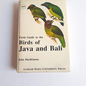 Può includere: Un libro con copertina flessibile intitolato Field Guide to the Birds of Java and Bali di John MacKinnon, pubblicato da Gadjah Mada University Press. La copertina color bianco sporco presenta tre illustrazioni colorate di uccelli su rami e un logo UGM.