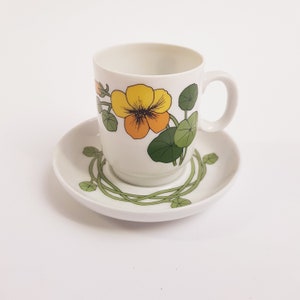 Pode incluir: Conjunto de chávena e pires de café em cerâmica branca. A chávena apresenta um desenho de flor amarela e laranja com folhas verdes. O pires tem um padrão de videira verde na borda. A chávena tem uma pega.