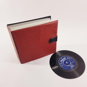 May include: A red textured vinyl record storage album with a black snap closure and a black spine, accompanied by a single black 45 RPM vinyl record featuring a blue Philips label with white text, including the brand name and record details.