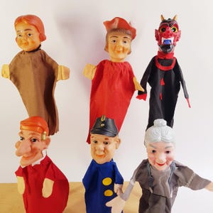 Könnte beinhalten: Eine Sammlung von sechs Handpuppen mit verschiedenen Charakteren, darunter eine Figur mit roten Haaren in braunem Gewand, eine Figur mit rotem Hut und Tunika, eine rote Teufelsfigur, eine Puppe mit großer Nase in Rot, eine Figur in blauer Uniform und eine ältere Person.