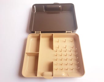 May include: An open, two-toned plastic craft box. The lid is dark brown, and the interior is beige. The box has compartments and pegs for organizing small items. The box is designed to hold sewing supplies, beads, or other craft materials.