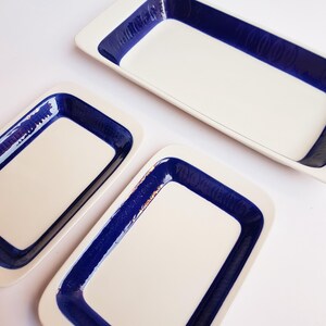 Pode incluir: Um conjunto de três travessas retangulares. As travessas têm interior branco e uma borda azul escura. A borda azul apresenta um design texturizado. As travessas estão dispostas sobre uma superfície branca.