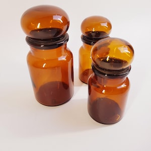 May include: Three amber glass apothecary jars with rounded lids. The jars have a cylindrical body and a dark brown base. The lids are a lighter amber color and sit atop a black rim. The jars are arranged on a white surface.