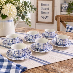 Könnte beinhalten: Ein Set aus fünf weißen Keramik-Teetassen mit passenden Untertassen, jeweils verziert mit einem blauen Blumen- und Geometriemuster. Die Tassen haben einen geschwungenen Henkel und einen breiten Rand und stehen auf einem Tisch mit blau-weiß gestreiftem Läufer.