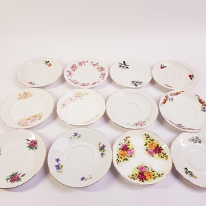 Op de afbeelding: Een verzameling vintage porseleinen schotels. Elke schotel heeft een uniek bloemmotief in verschillende kleuren, waaronder roze, rood, geel en paars, met gouden rand. De schotels zijn gerangschikt op een witte ondergrond.