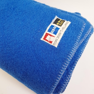 Pode incluir: Uma manta de lã azul vibrante com uma superfície texturizada e uma borda costurada. Uma etiqueta retangular branca está anexada, apresentando um ícone de gato vermelho, o texto DIDAS DEKEN HOLLAND e uma seção preta com três estrelas amarelas.