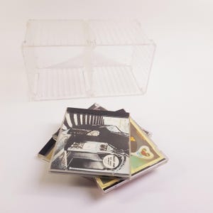 May include: A clear, plastic CD storage unit with a two-compartment design. Several CD cases are stacked below. The CD cases feature album art in black, white, and yellow. One CD case has a sticker that reads "MAZEY STAR".