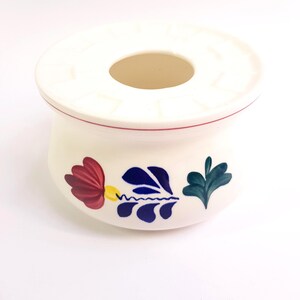 Op de afbeelding: Een witte keramische asbak met een bloemmotief. De asbak heeft een ronde bovenkant met een centrale opening en is versierd met rood, blauw en groen geschilderde bloemen. Een dunne rode lijn omcirkelt de basis van de bovenkant.