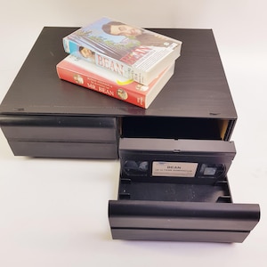 Può includere: Un mobiletto porta cassette VHS in plastica nera con un cassetto aperto contenente una cassetta VHS nera con l'etichetta Bean De Ultieme Rampenfilm. Due libri intitolati Bean e Mr. Bean sono impilati sulla parte chiusa del mobiletto.