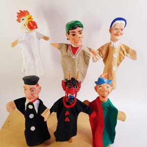 Op de afbeelding: Een verzameling van zes handpoppen met diverse personages, waaronder een haan, een persoon met een groene pet, een ouder figuur, een persoon met een zwarte pet, een rode duivel met hoorns en een persoon met een blauwe hoed, allen met stoffen lichamen.