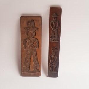 Può includere: Due stampini per biscotti in legno vintage, uno con una donna con un cappello e l'altro con un uomo e una donna con cappelli.
