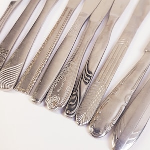 Può includere: Una collezione di coltelli d'argento vintage con vari disegni del manico. I coltelli sono disposti diagonalmente, mostrando diversi motivi, tra cui dettagli floreali, geometrici e testurizzati. Le lame sono lucenti, riflettendo la luce.