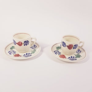 Könnte beinhalten: Zwei weiße Kaffeetassen aus Keramik mit blauen und roten Blumendesigns an der Seite. Jede Tasse steht auf einer passenden Untertasse mit dem gleichen Blumendesign.