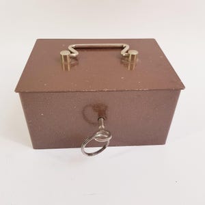 Op de afbeelding: Een rechthoekige bruine metalen afsluitbare doos met een gespikkelde afwerking. De doos is voorzien van een zilverkleurig metalen handvat op het deksel en een zilverkleurige sleutel in het voorste slot, met een ring aan de kop van de sleutel.