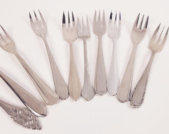 May include: A collection of vintage silver forks, each with a unique handle design. The forks vary in size and style, with tines ranging from three to four. The arrangement showcases the variety of silverware designs.